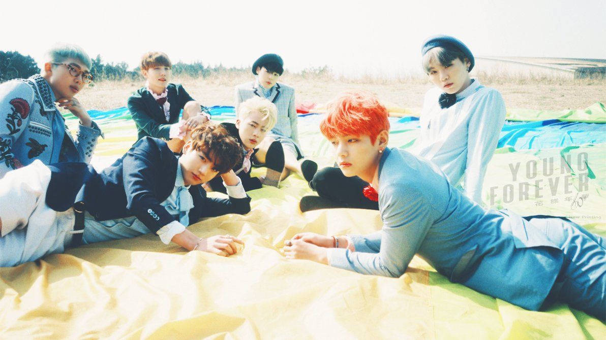 Miembros de BTS recostados sobre una cobija en la arena de la playa durante el verano.