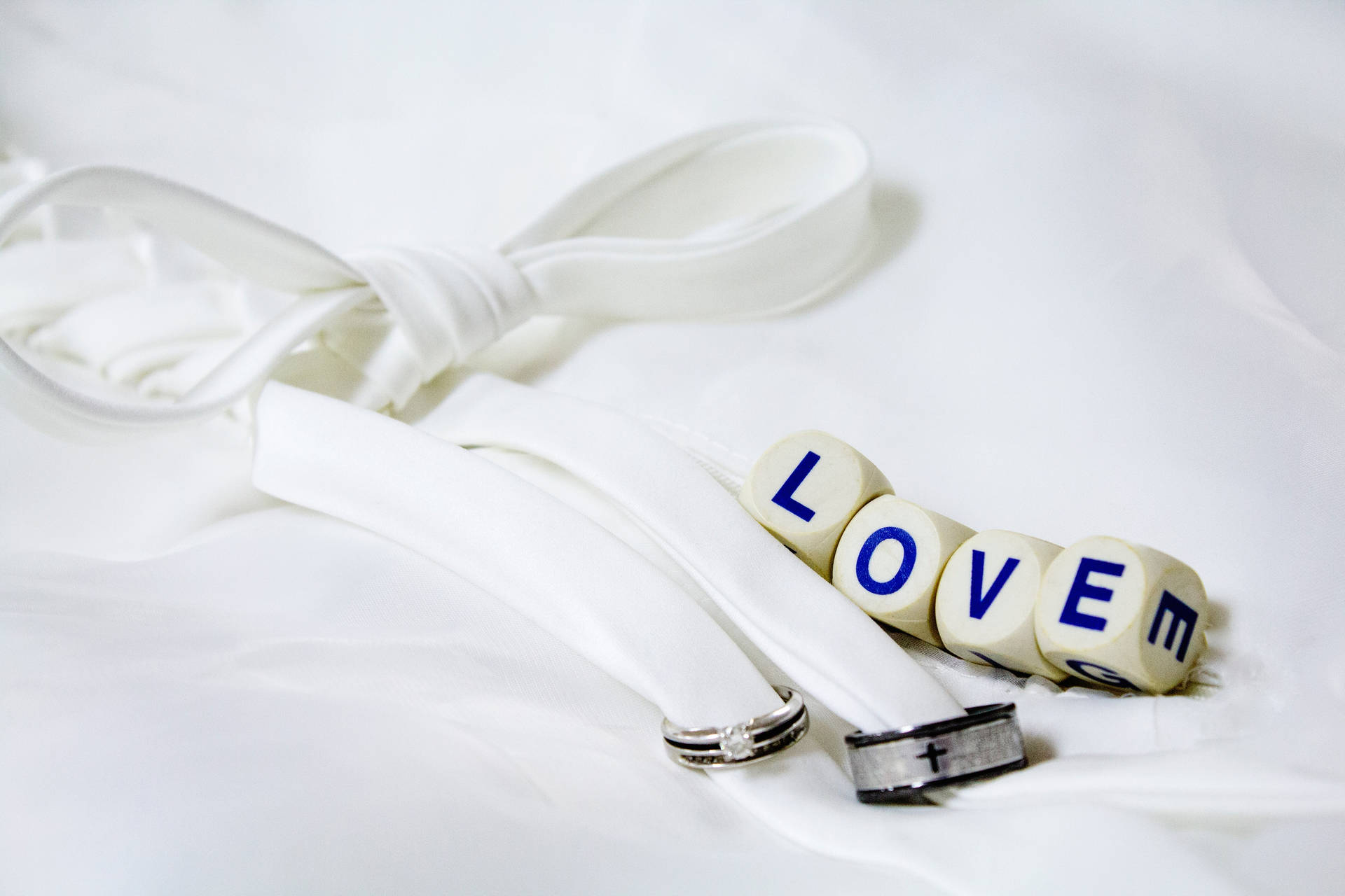 Escrito "Amor" en cuatro dados blancos junto a dos anillos de boda plateados y negros atados con un listón blanco sobre una sábana blanca.