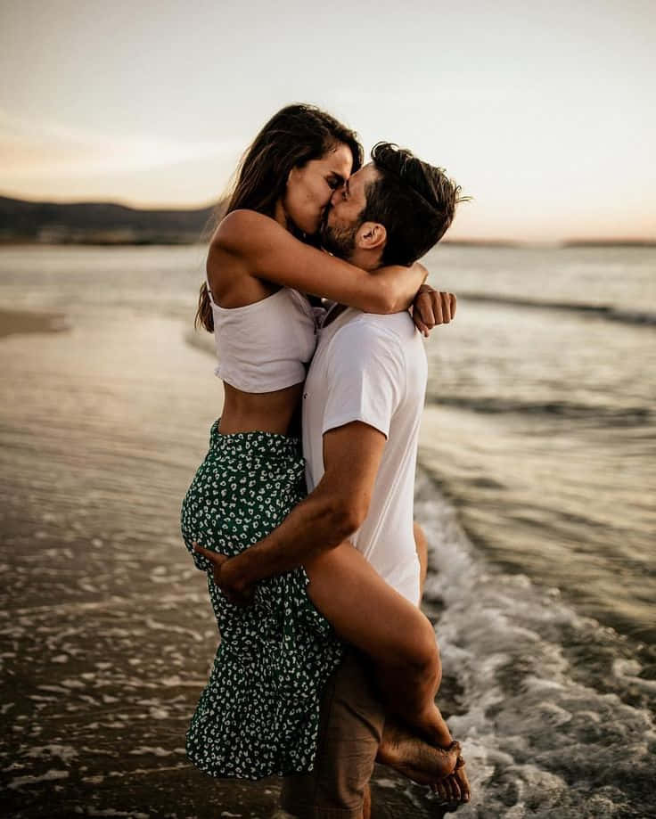 Pareja en la playa besándose mientras él la carga entre las olas Fondos de pantalla