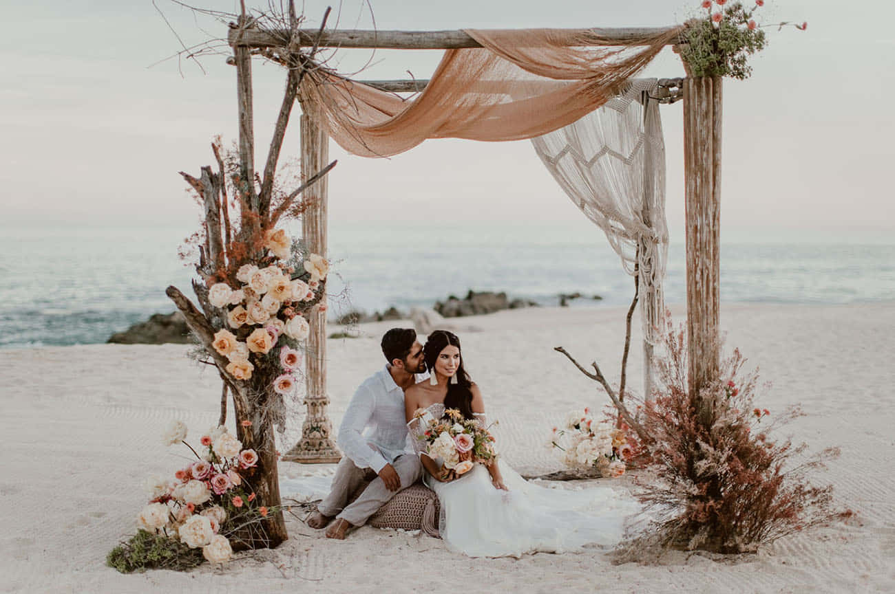 una novia y un novio sentados en un arco de madera en la playa Fondos de pantalla