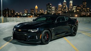 Fondo de pantalla de Un Camaro ZL1 negro estacionado en un estacionamiento en la azotea con el horizonte de la ciudad de noche.