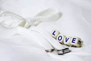 Escrito "Amor" en cuatro dados blancos junto a dos anillos de boda plateados y negros atados con un listón blanco sobre una sábana blanca.