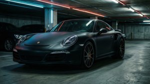 Fondo de pantalla de Un Porsche 911 Turbo S en negro mate destaca en un garaje subterráneo ambiental con reflejos de neón.