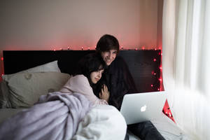 Pareja en la cama usando un MacBook Air junto a una ventana con cortina blanca. — MacBook Pro Fondos de pantalla
