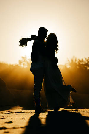 Silueta de pareja de novios enamorados abrazándose con la chica llevando un ramo de flores contra el atardecer y el cielo naranja amarillento.